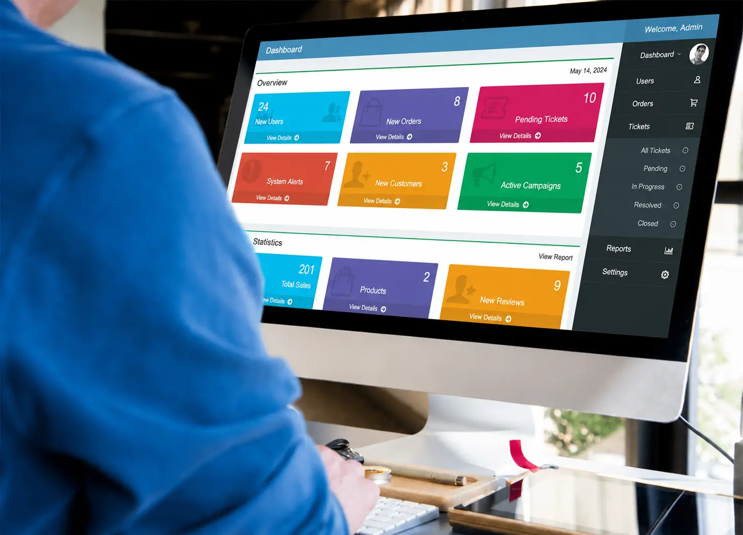 Person working at a desktop computer displaying a colorful English admin dashboard with overview cards, statistics, reports, and system management menu.