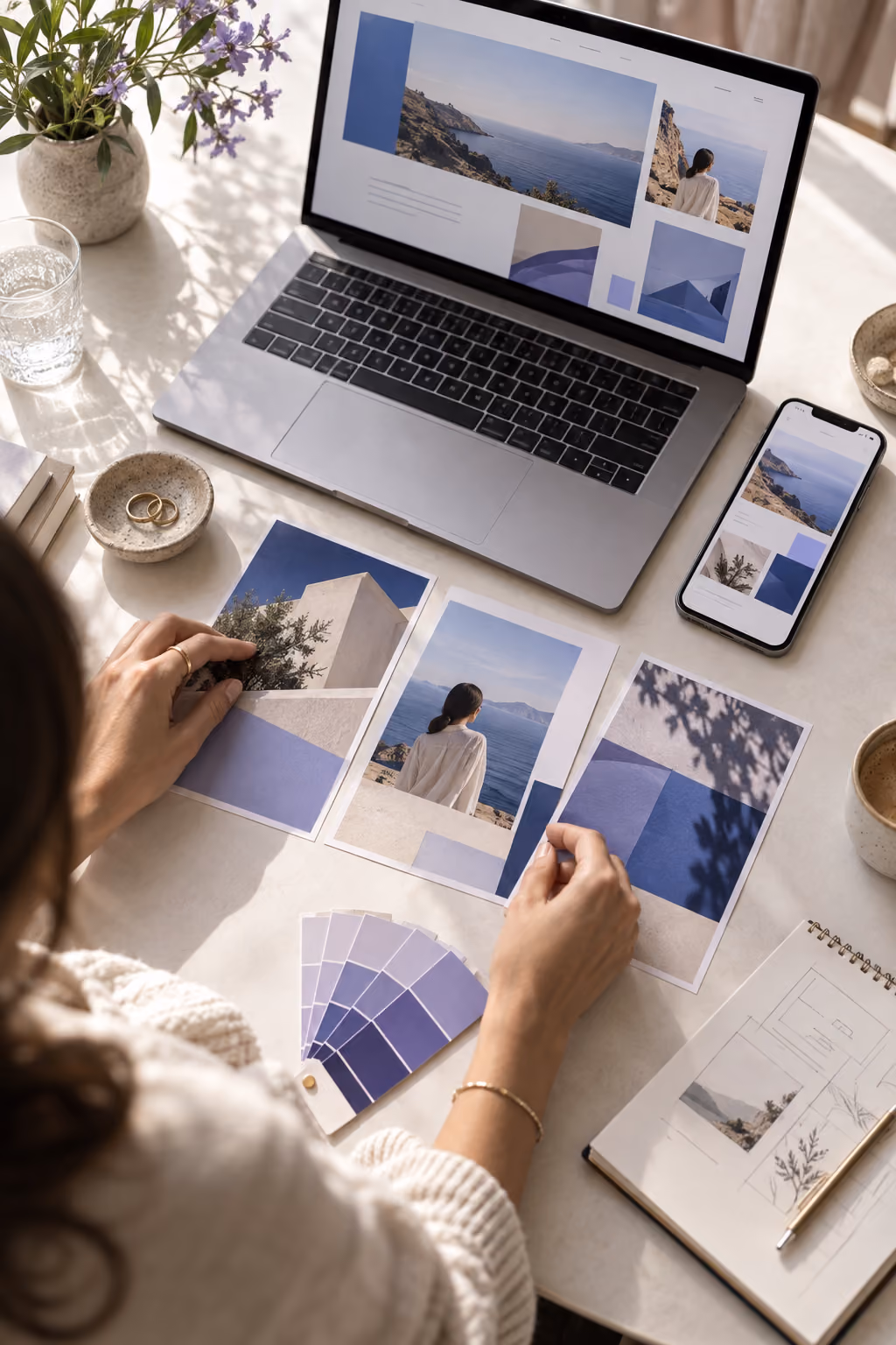 Person arranging printed photos of a coastal landscape with blue and beige tones on a white table, with a laptop and smartphone showing matching images, a color swatch book, a notebook with sketches, a cup of coffee, a glass of water, and a vase with purple flowers.