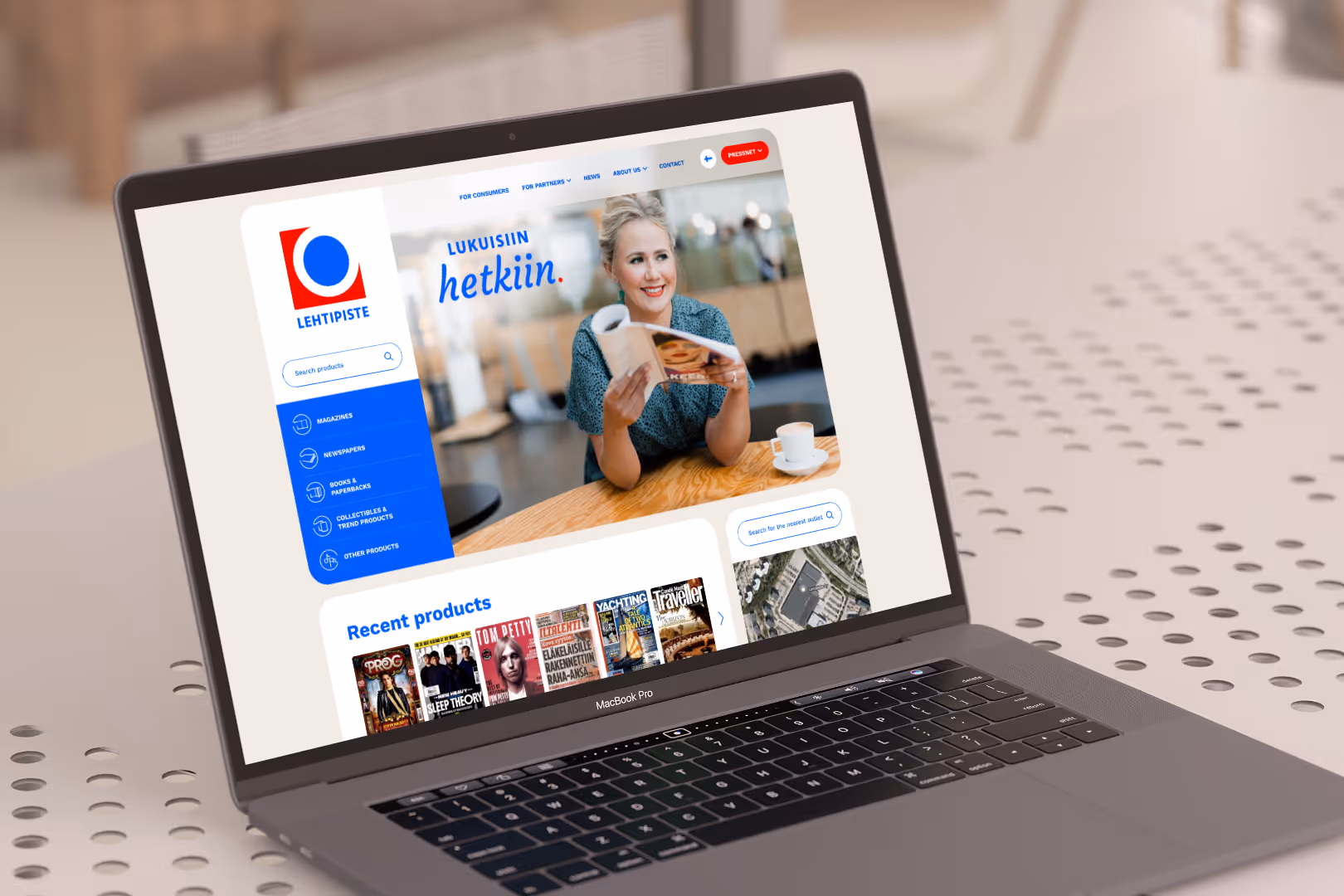 MacBook Pro, jossa näkyy Lehtipiste-verkkosivusto, johon sisältyy kuva hymyilevästä naisesta, joka lukee lehteä kahvipöydässä.