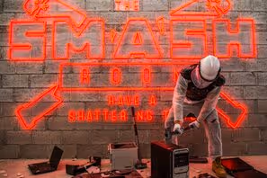 Person in safety gear smashing electronics with a sledgehammer inside The Smash Room.  
