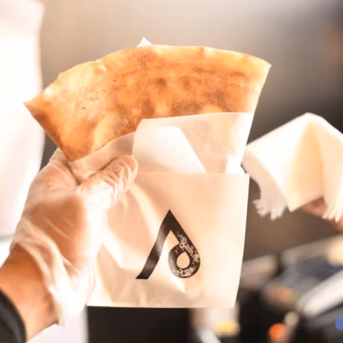 Hand holding a freshly made stuffed pastry in branded paper wrap.  
