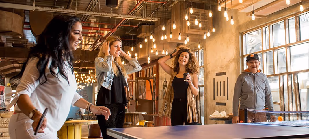 Group of friends playing ping pong and enjoying drinks in a modern entertainment venue with hanging lights.  
