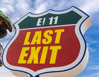 Large E11 Last Exit road sign against a blue sky.  
