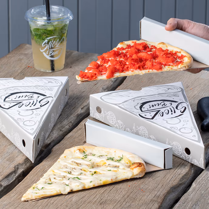 Slices of pizza served in branded triangular pizza boxes with a cold drink on a wooden table.  
