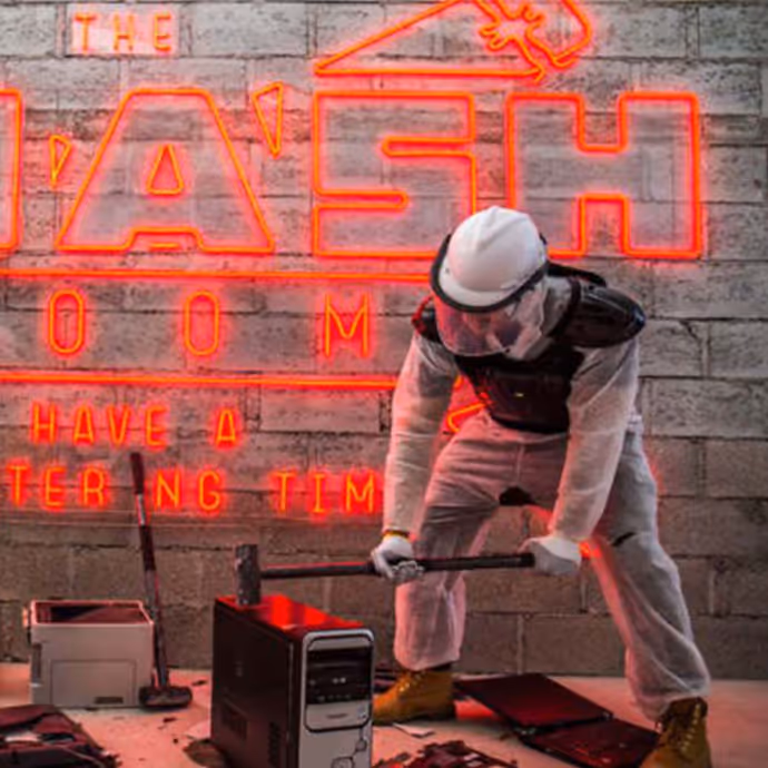 Person in protective gear smashing objects with a sledgehammer inside a rage room.  

