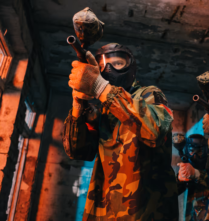 Person in camouflage gear playing paintball indoors with a paintball marker.  
