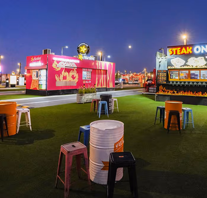 Colorful nighttime view of an outdoor food court with container-style eateries including Hardee's and Steak On, with artificial grass, barrel tables, and scattered stools.