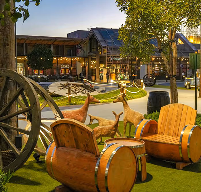 Charming outdoor setting with wooden barrel-style chairs, deer sculptures, wagon wheels, and ambient string lights in a village-themed area.