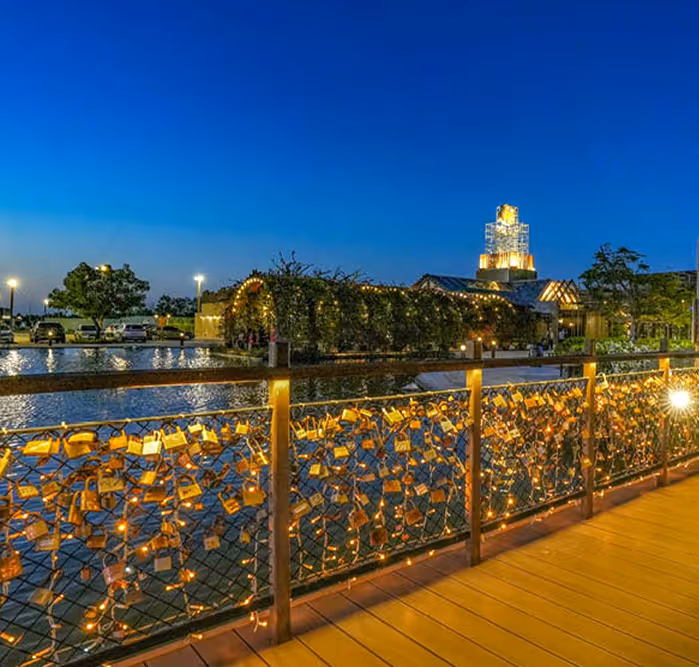 Scenic view of a love lock bridge decorated with fairy lights and padlocks at night, overlooking a tranquil water body and a building wrapped in greenery.