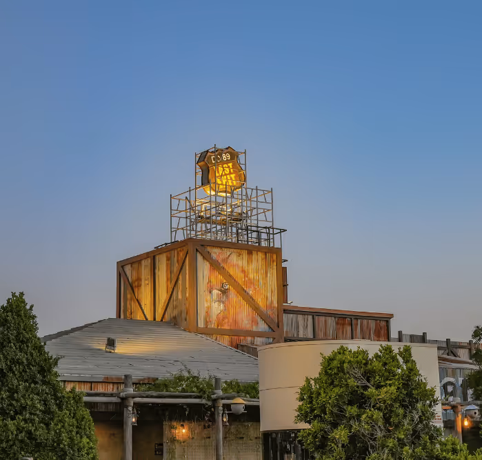 Rustic wooden building with a weathered exterior and a glowing "Last Exit" sign on top, surrounded by greenery at dusk.
