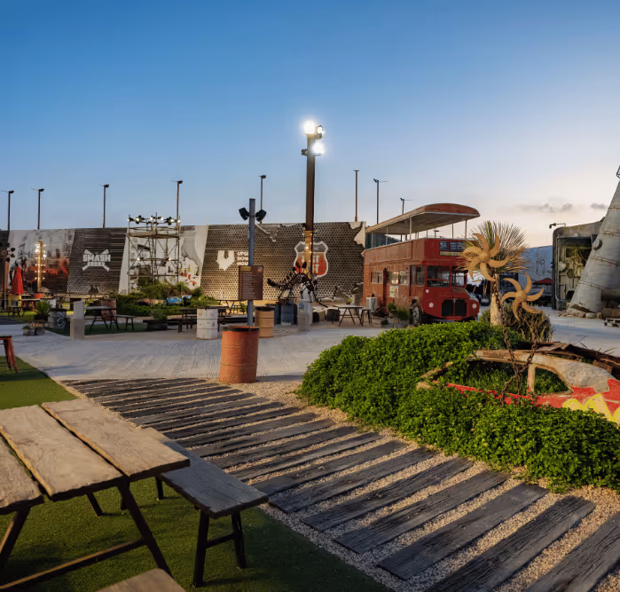 Eclectic outdoor space featuring picnic benches, artistic sculptures, plants, and a red double-decker bus under evening lights.