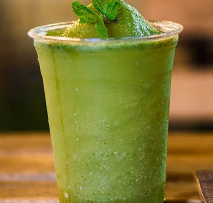 Refreshing green frozen drink garnished with mint leaves, served in a clear plastic cup on a wooden surface.