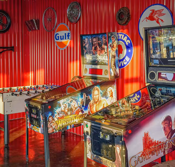 Retro-style arcade area featuring pinball machines themed after WWE WrestleMania and Game of Thrones, along with a foosball table and vintage car-themed wall decor.
