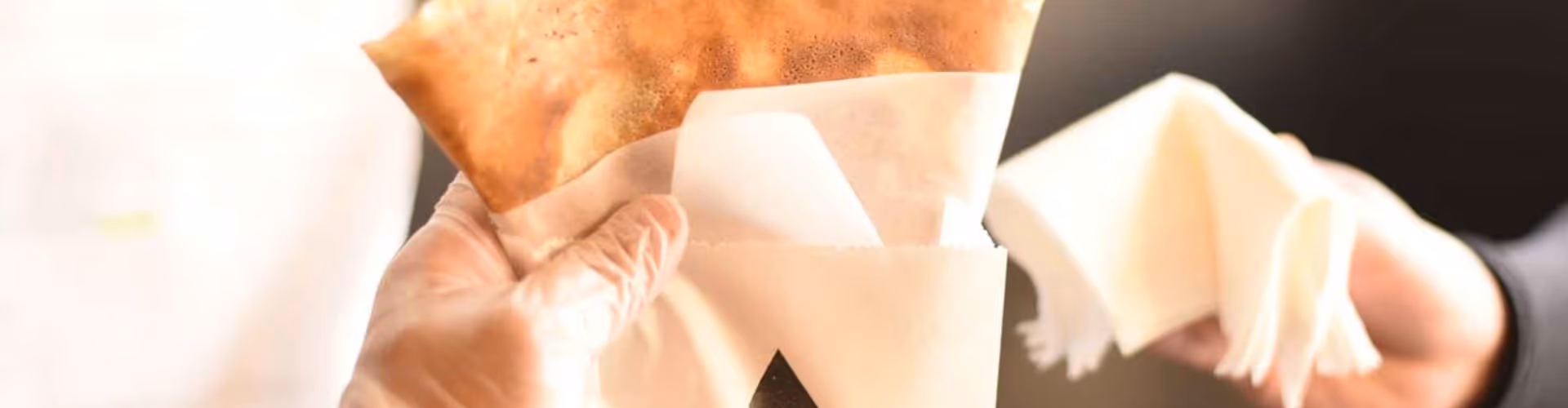 gloved hand holding a crispy, golden-brown folded pastry in white paper