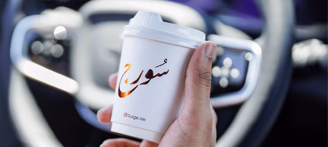 a hand holding a white takeaway coffee cup with arabic script, inside a car.