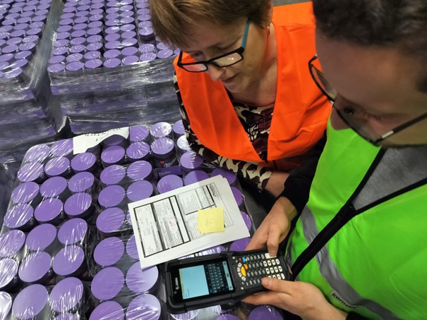 Two warehouse workers in orange and green safety vests reviewing inventory with a handheld scanner and paperwork next to pallets of purple-lidded containers.