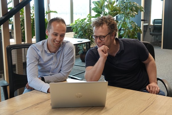 Two men sitting at a wooden table in an office, smiling and looking at a laptop screen.