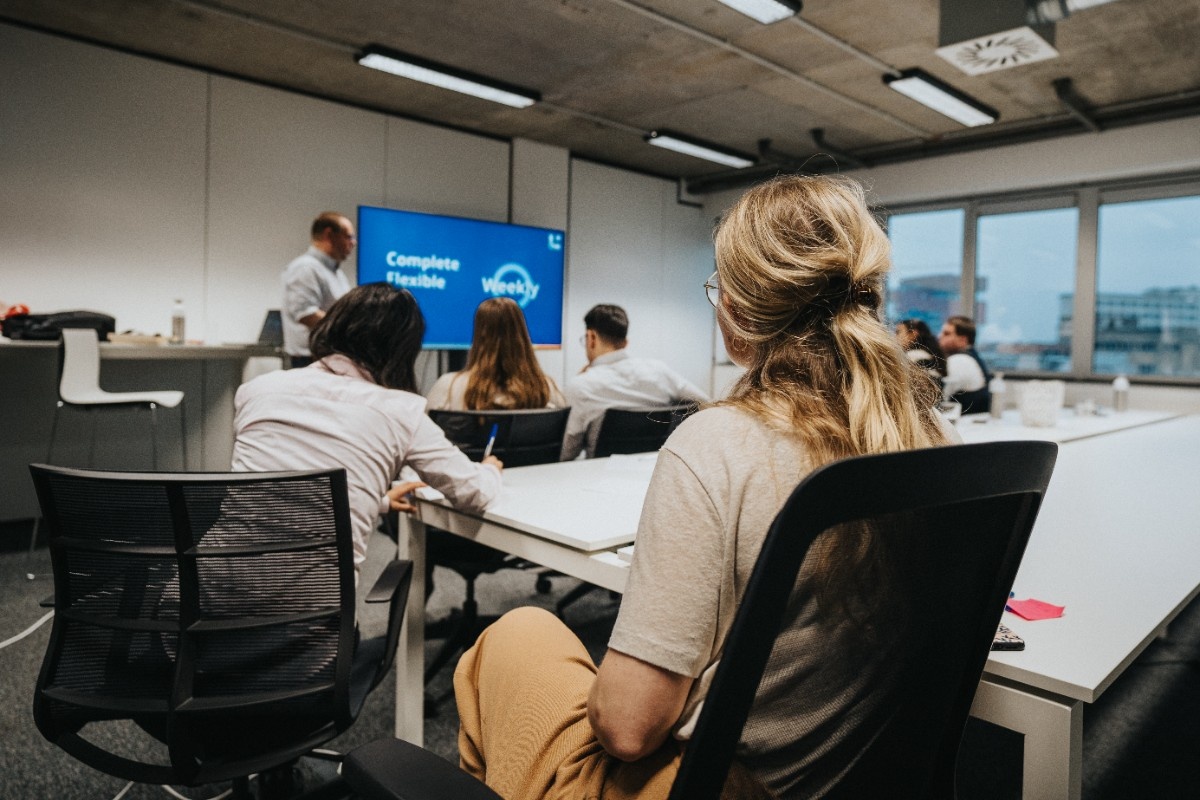 People seated around a conference table listening to a man giving a presentation with a blue screen in the background.
