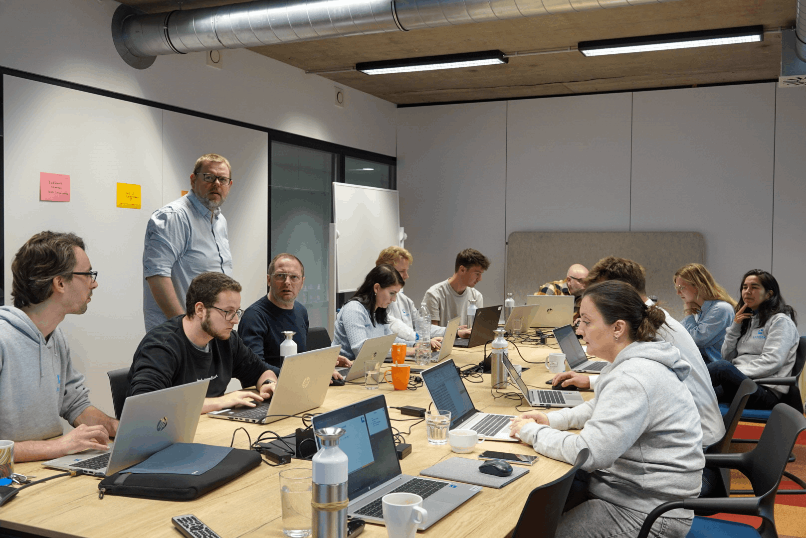 A group of eleven people in a meeting room working on laptops around a large table with water bottles and coffee cups.