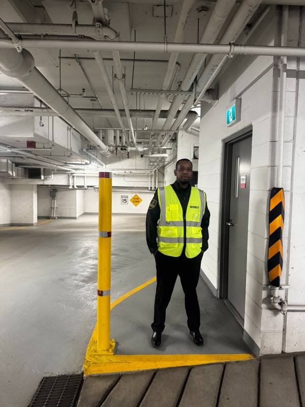 Security guard wearing a neon yellow safety vest standing inside a parking garage near a door with an exit sign above it.