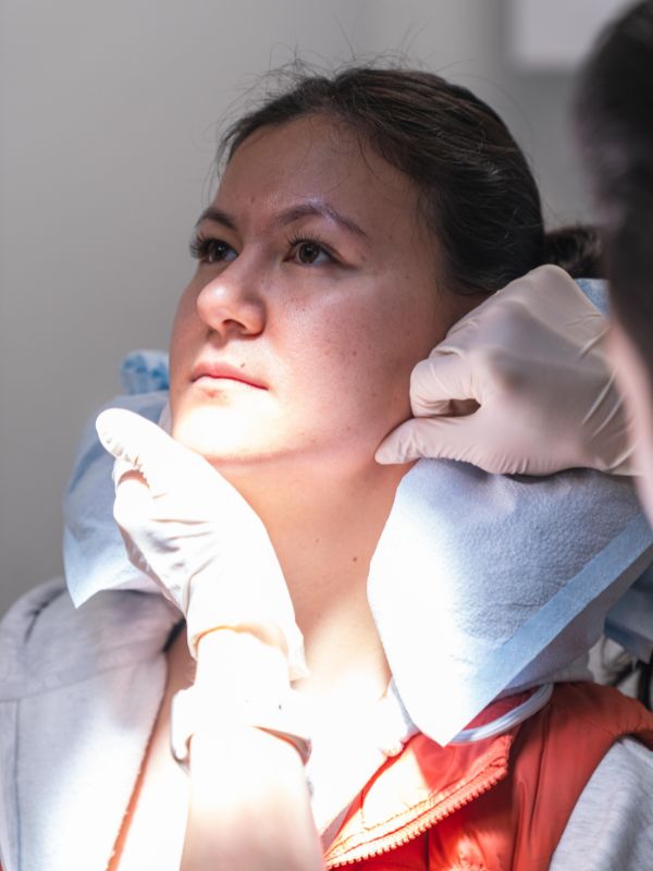 A woman receiving a facial examination with hands in gloves gently touching her jawline, under bright clinical lighting.