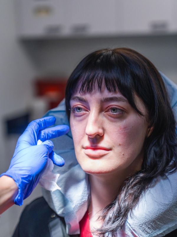 A woman with dark hair sits wrapped in a light blue blanket as a gloved hand gently touches her face.