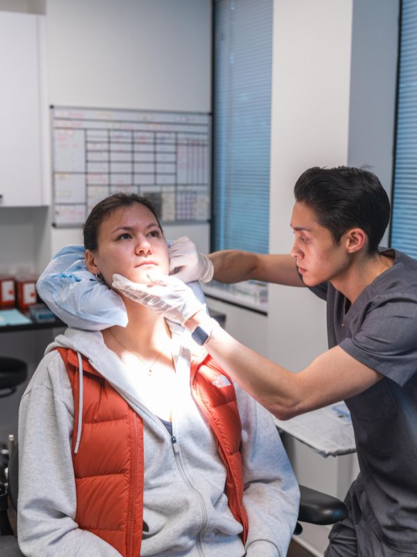 Medical professional wearing gloves examining a woman's face under focused light in a clinical setting.