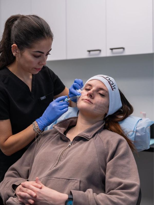 Woman receiving a cosmetic injection in her cheek from a clinician wearing blue gloves in a medical setting.