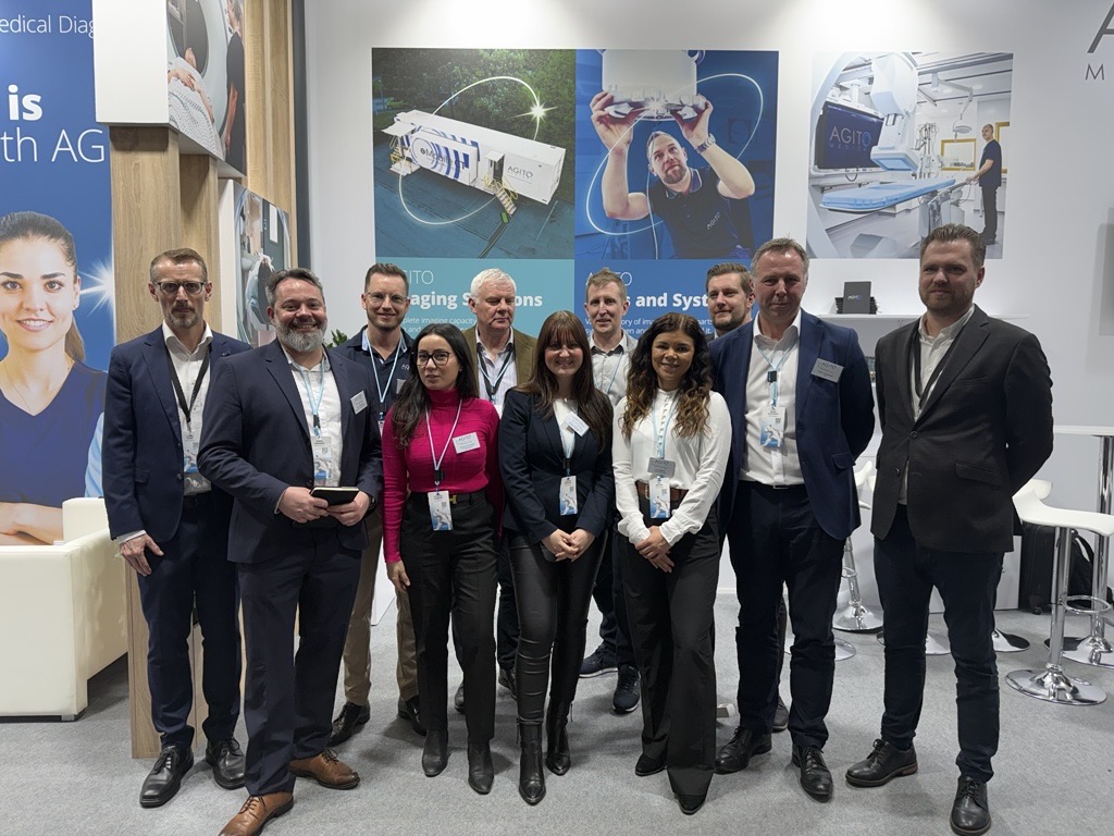 Group of eleven professionally dressed people posing at a medical technology exhibition booth with posters and equipment in the background.