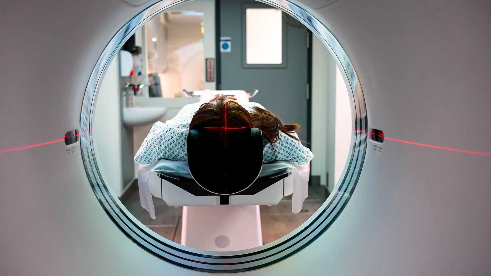 Patient lying face down on a medical scanner table with red laser alignment lights on head inside a CT or MRI machine.