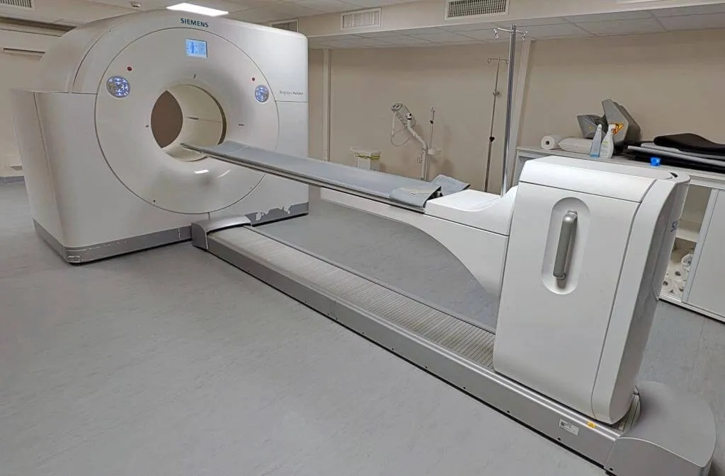 Siemens medical imaging machine in a clinical room with patient bed and surrounding medical equipment.