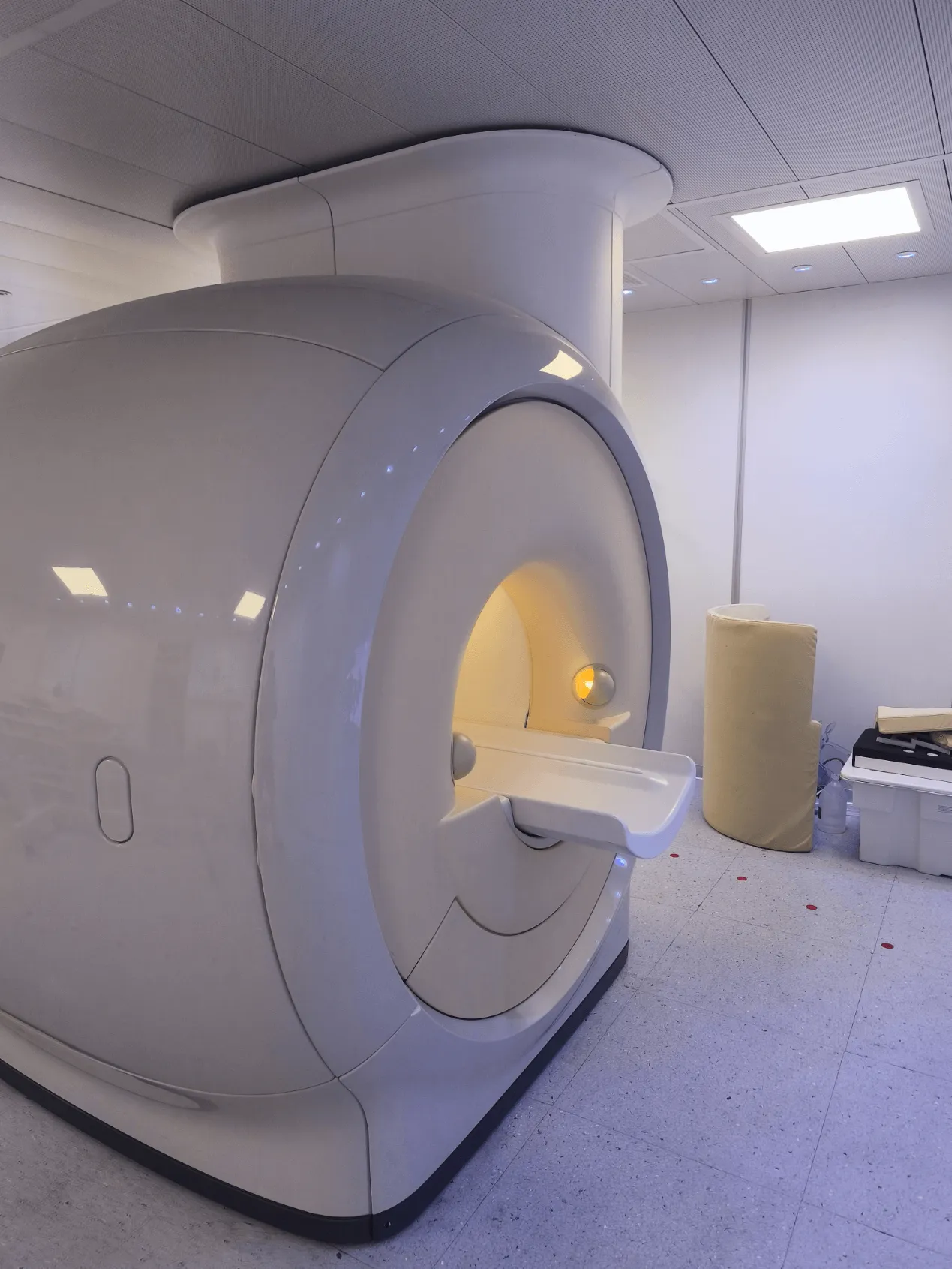 Modern MRI machine in a clean, well-lit medical examination room with a padded cylindrical object and storage boxes nearby.