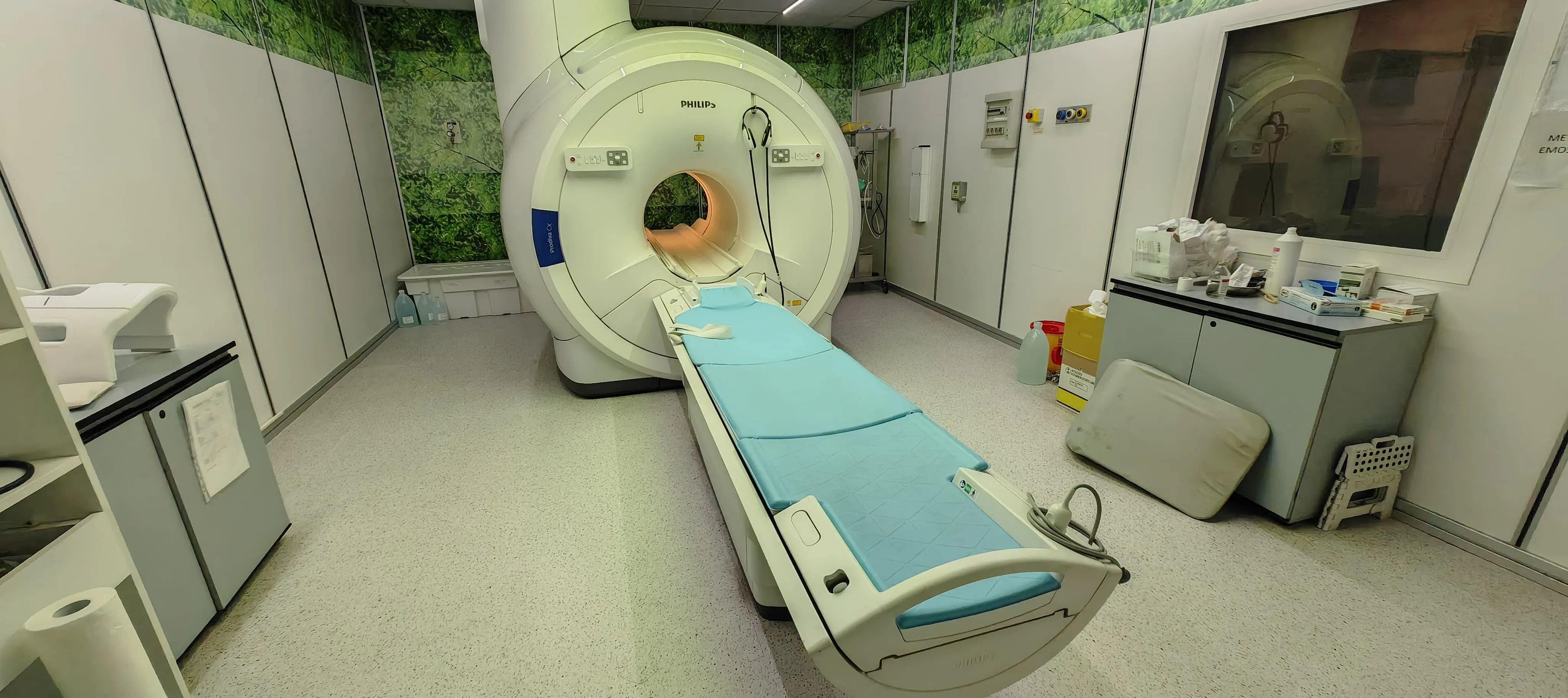 Philips MRI machine with a patient table covered in blue padding inside a medical imaging room with cabinets and medical supplies.