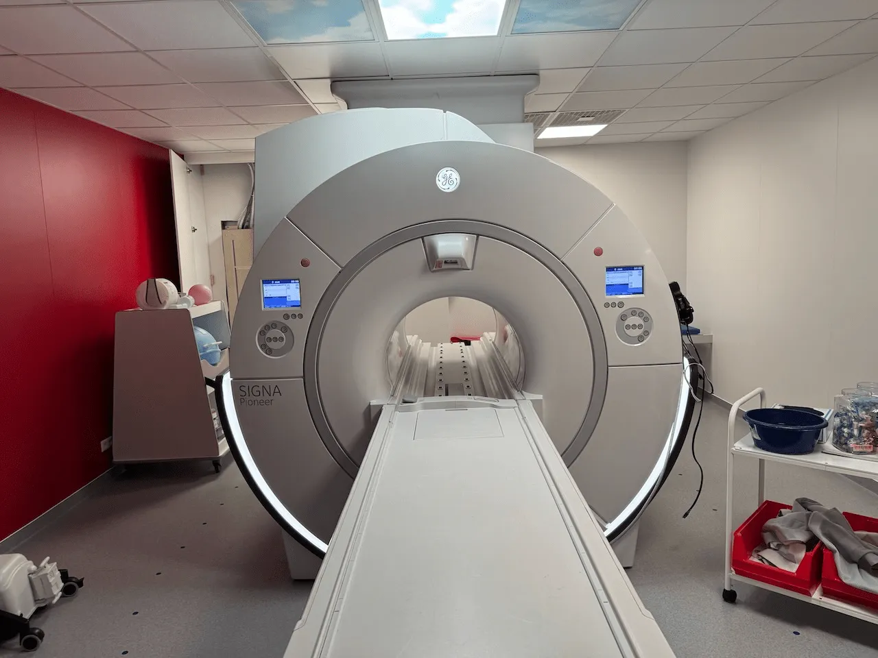 MRI machine with patient table extending towards the viewer inside a medical room with red and white walls.