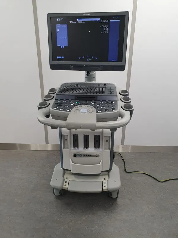 Siemens medical ultrasound machine with a monitor, keyboard, control panel, and wheels, placed on a gray floor.