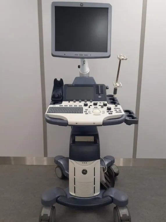 Medical ultrasound machine with a large monitor, control panel, and wheels, positioned in a clinical setting.