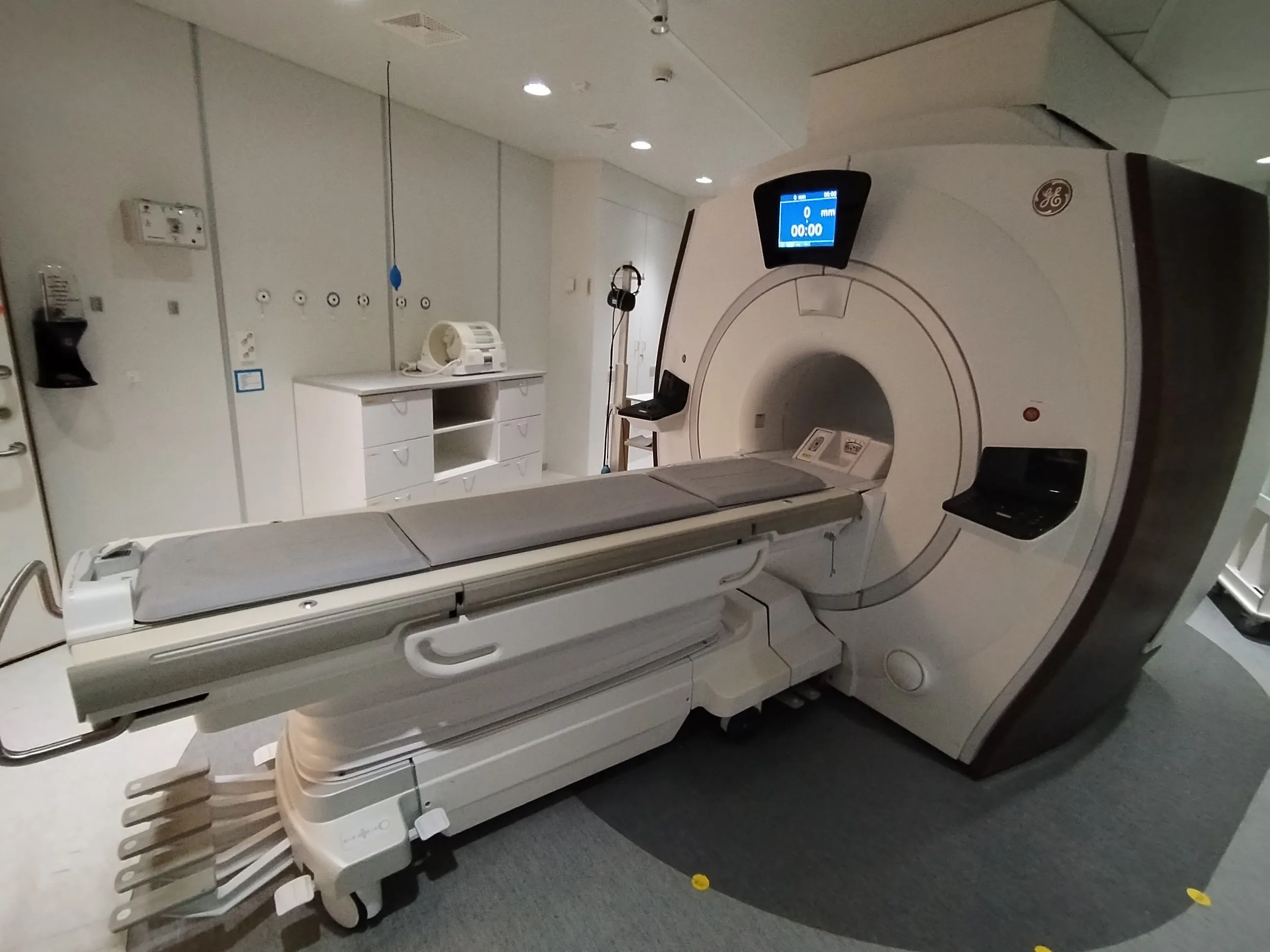 Interior view of a medical room featuring a large white GE MRI machine with an empty patient bed and attached control panel.