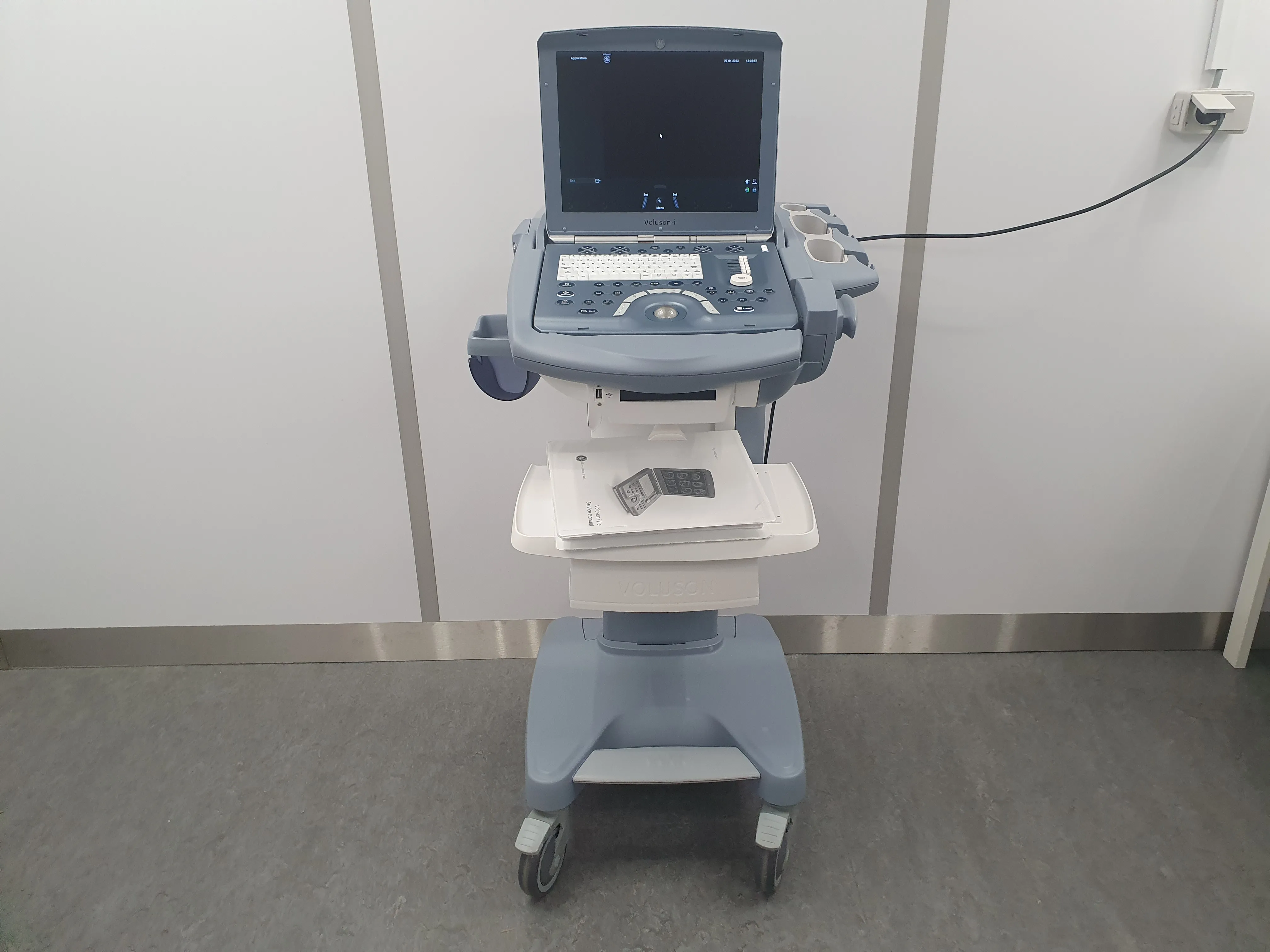 Medical ultrasound machine with monitor, control panel, and wheels in a clinical room.