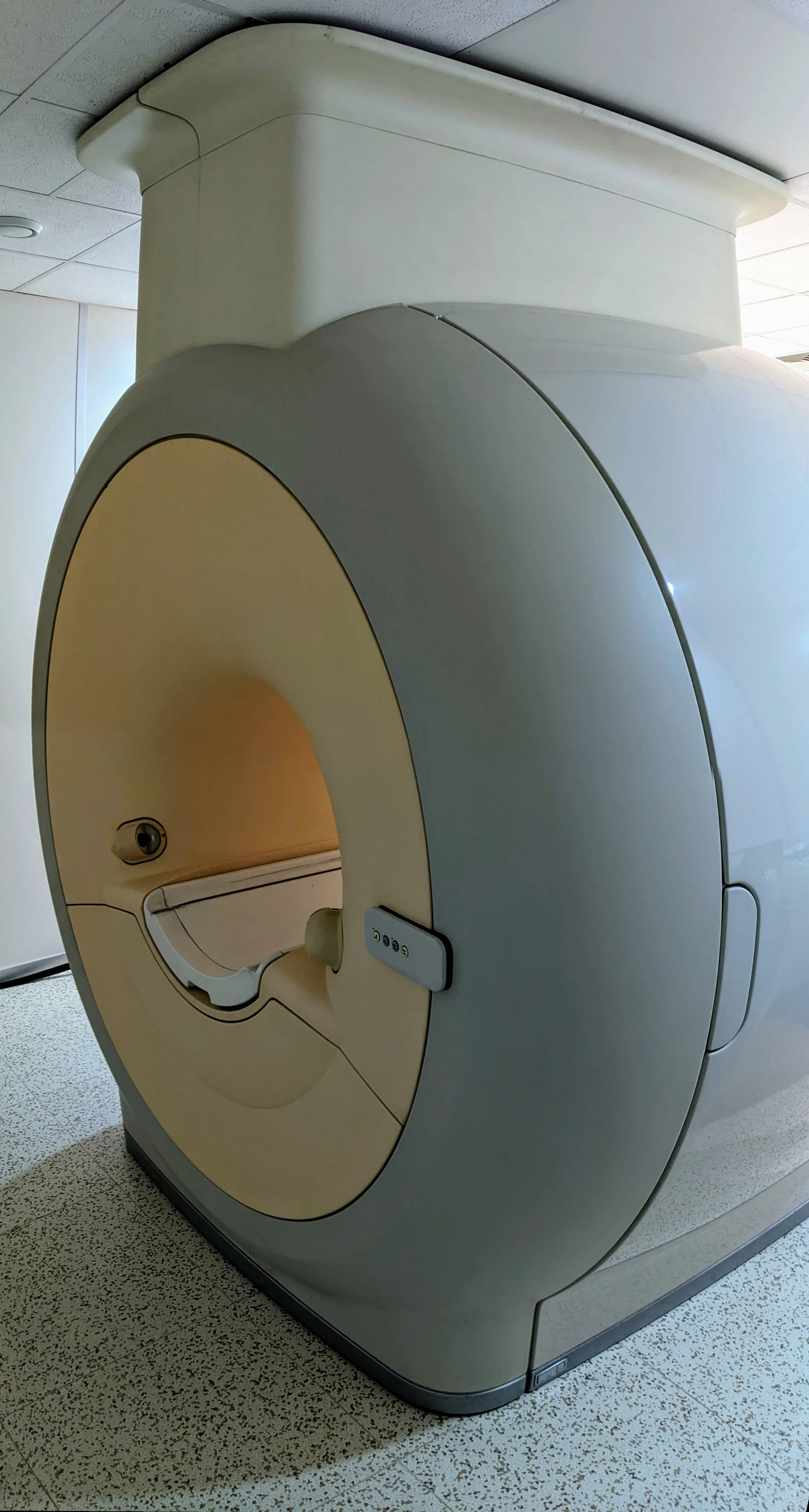 Side view of a large MRI scanner machine in a clinical room with speckled flooring and a white ceiling.