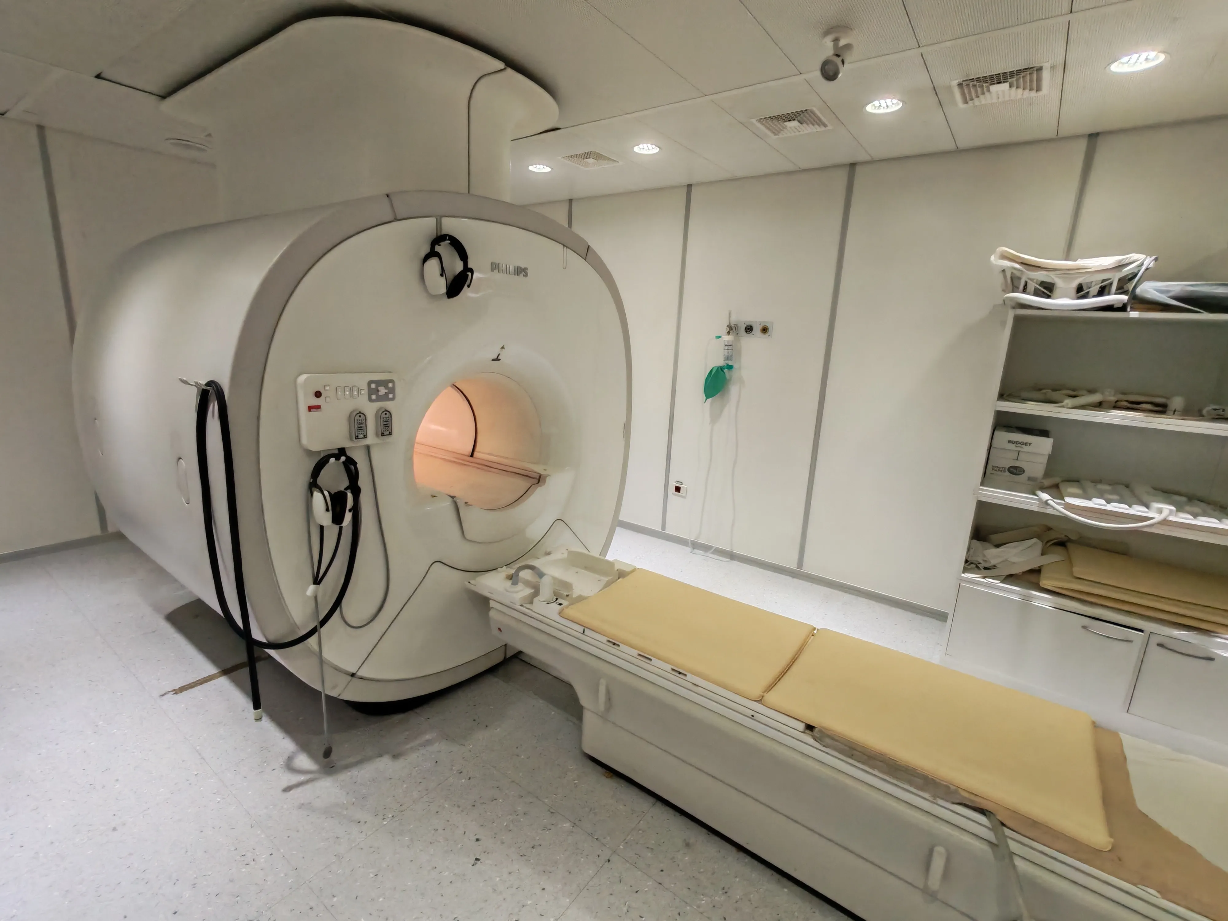 Philips MRI machine with patient table and headphones in a clinical room with shelves holding supplies.