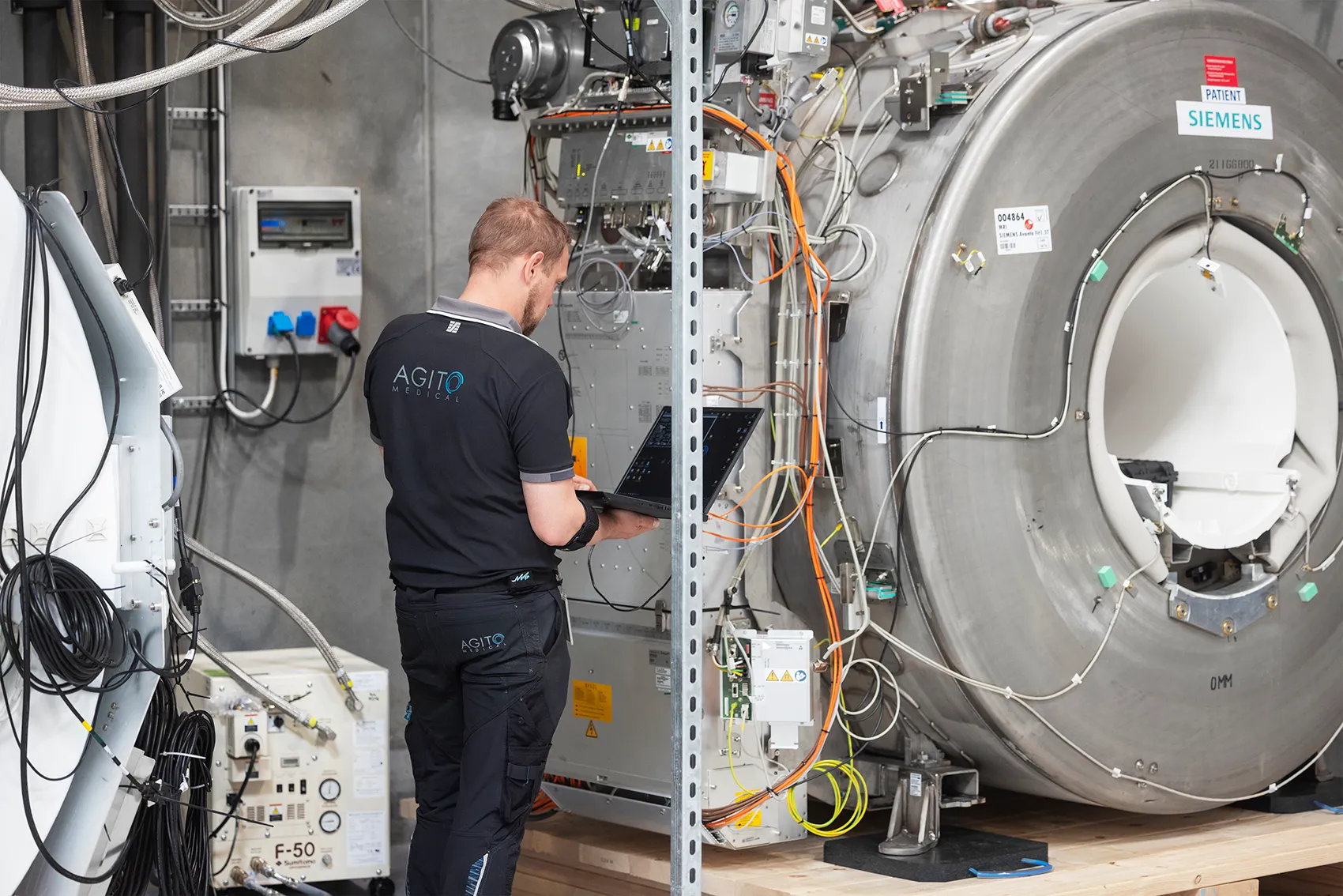 Technician in AGITO Medical uniform using a laptop to inspect and maintain a Siemens medical device in a technical room.
