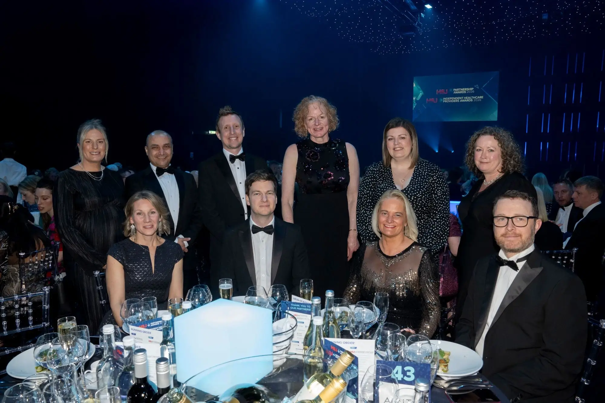 Group of men and women in formal wear seated and standing around a dinner table at an awards event.