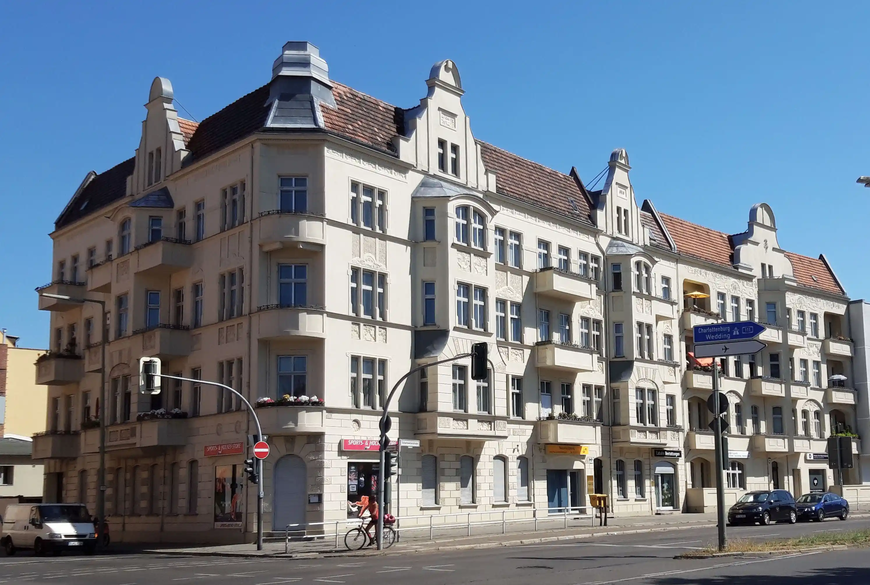 Mehrstöckiges Wohn- und Geschäftsgebäude mit hellen Fassaden, Balkonen und roten Ziegeldächern an einer Straßenecke bei klarem Himmel.