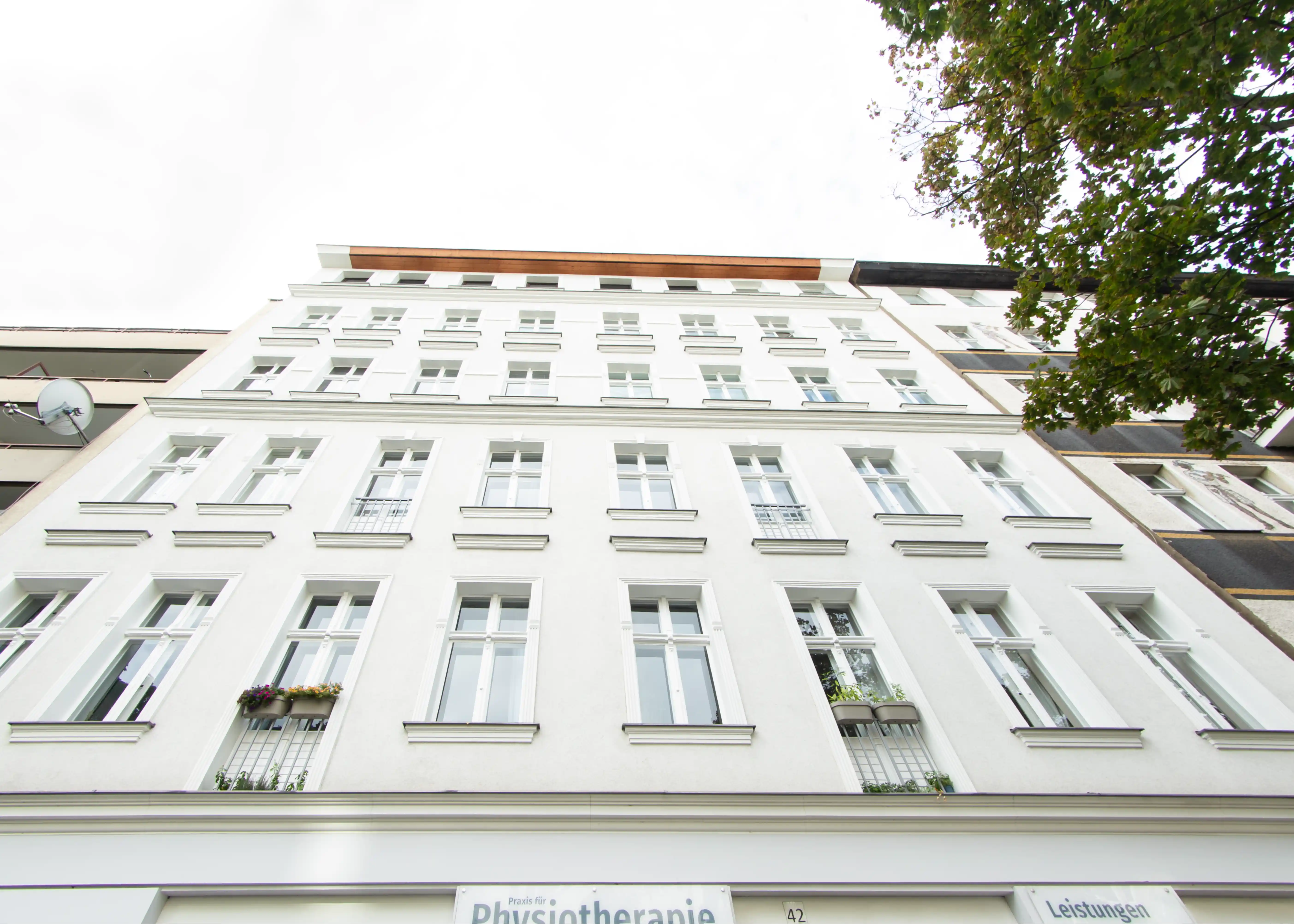 Blick nach oben auf eine weiße mehrstöckige Gebäudefassade mit vielen Fenstern und einigen Blumenkästen.