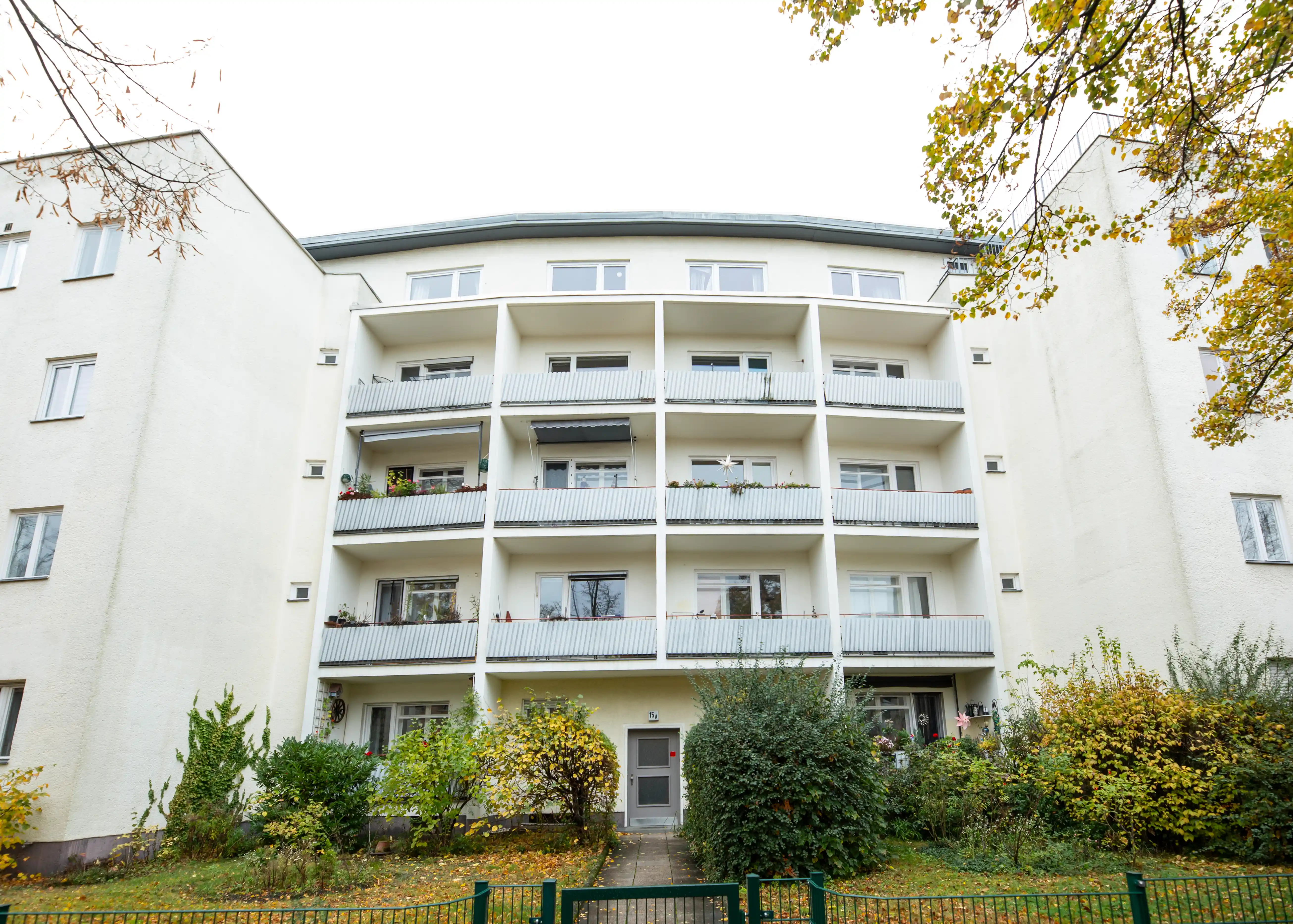 Vorderansicht eines mehrstöckigen Wohngebäudes mit Balkonen und herbstlichen Bäumen und Sträuchern im Vorgarten.