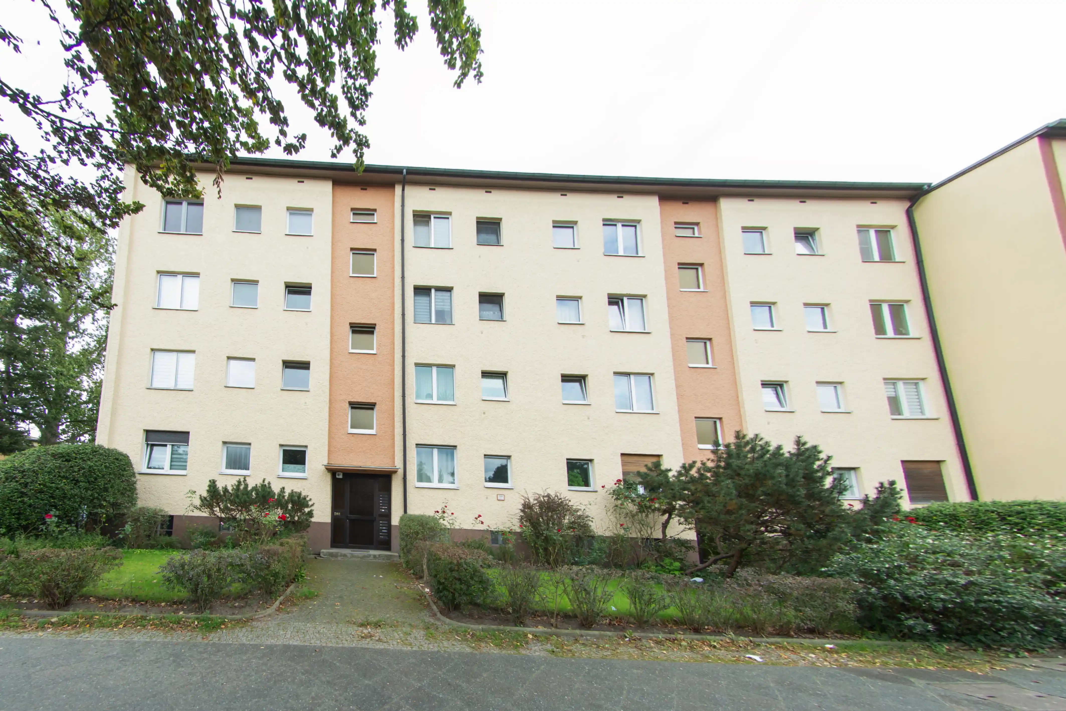 Mehrstöckiges Wohngebäude mit hellem Putz, mehreren Fenstern und grünem Vorgarten mit Büschen und Bäumen.