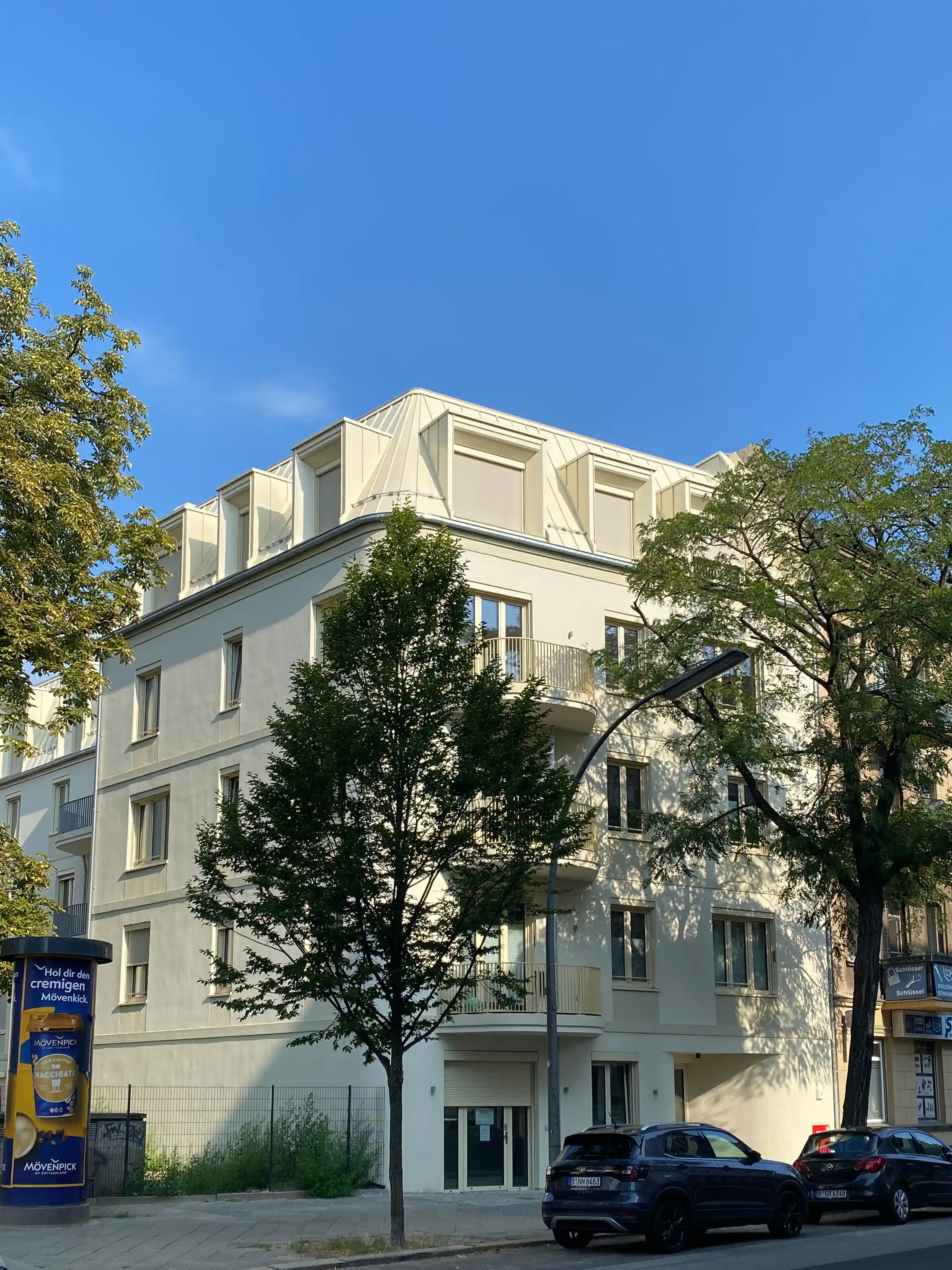 Modernes, vierstöckiges Wohngebäude an einer ruhigen Straße mit parkenden Autos und Bäumen bei blauem Himmel.