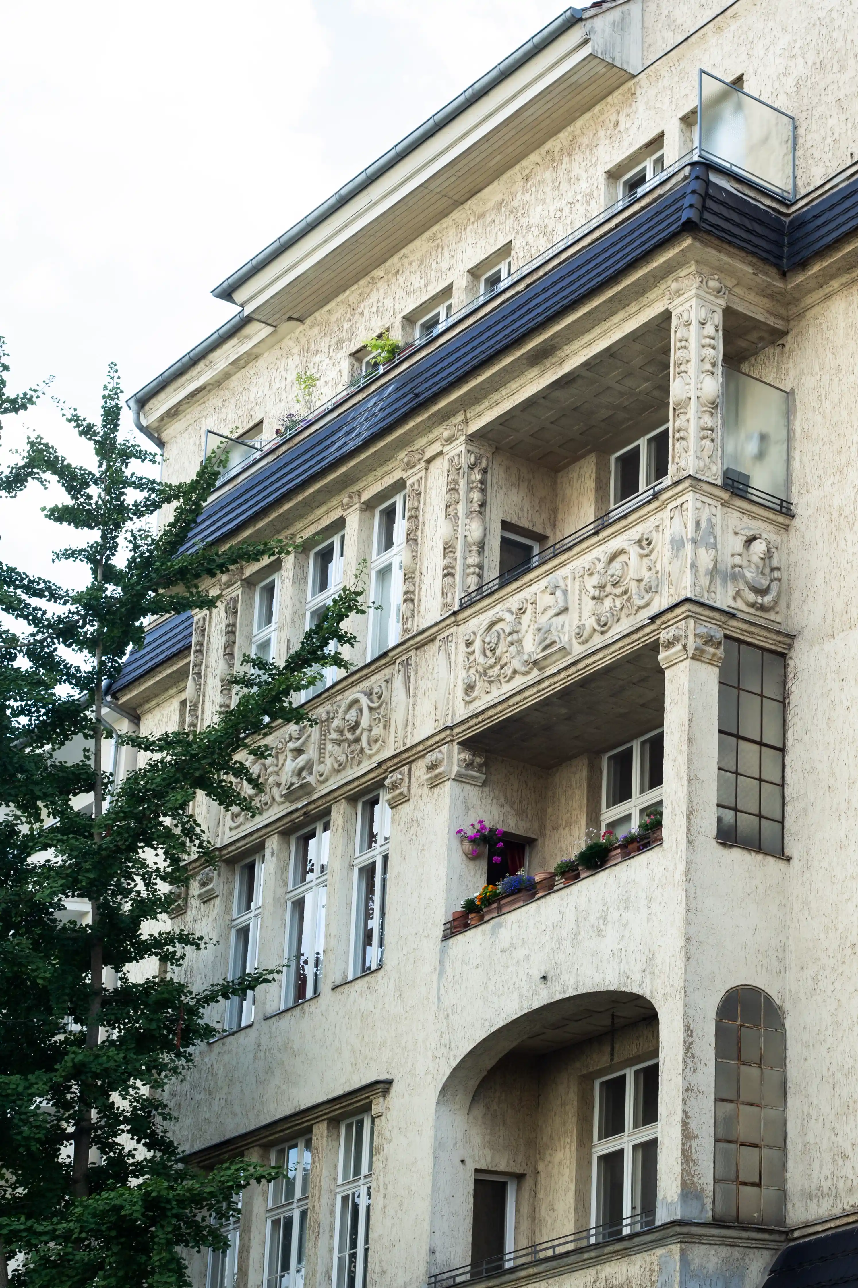 Fassade eines Altbaus mit verzierten Balkonen, Blumenkästen und einem Baum im Vordergrund.