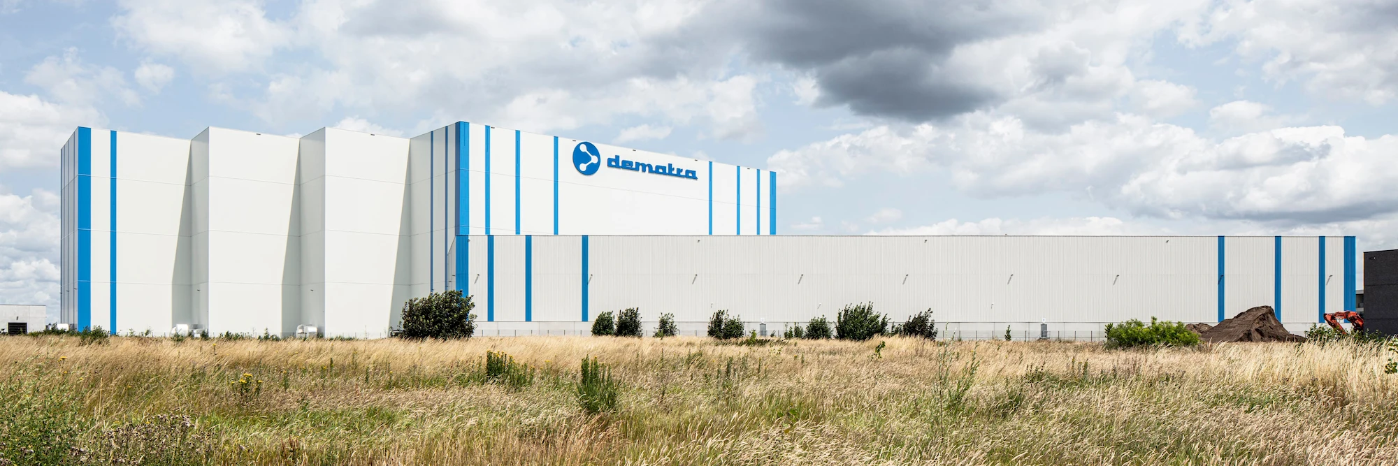 Large white industrial building with blue vertical stripes and Dematra logo, surrounded by tall grass under a cloudy sky.