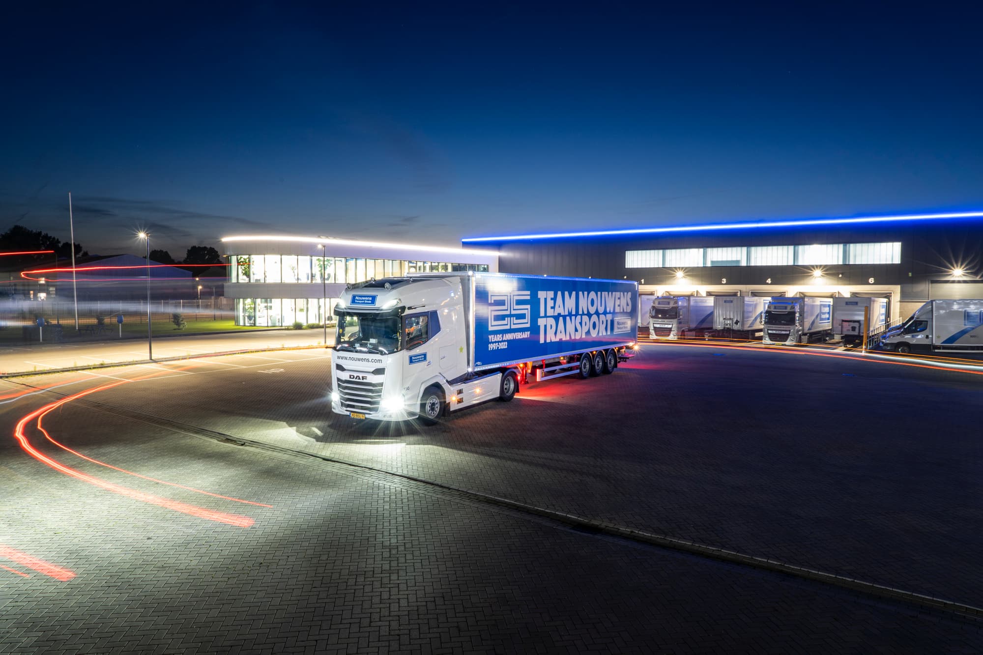 Illuminated white and blue Team Nouwens Transport truck at a loading dock area during twilight.